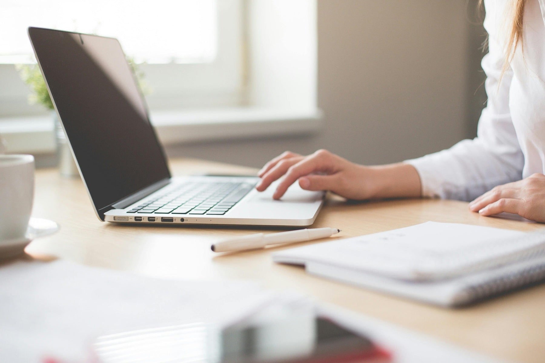 Person working on laptop at desk with notebook and coffee in bright home office